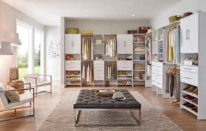 A spacious, well-organized walk-in closet with white cabinetry, multiple drawers, shelves, and hanging space, featuring neatly arranged clothes, shoes, and accessories. The room also has large windows, natural light, and a cozy seating area with modern chairs and a plush ottoman.