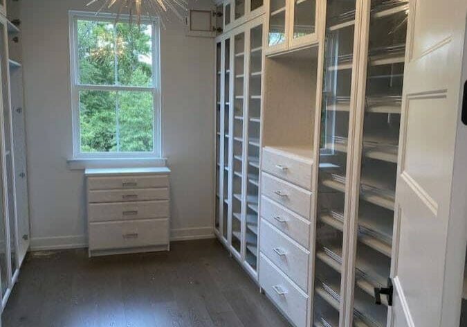 A spacious walk-in closet with white shelving units, drawers, and cabinets, featuring a modern chandelier and a large window letting in natural light.