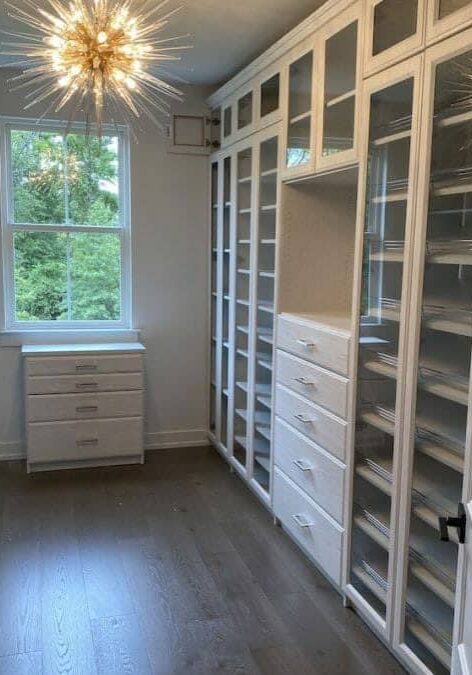 A spacious walk-in closet with white shelving units, drawers, and cabinets, featuring a modern chandelier and a large window letting in natural light.
